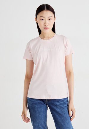 T-shirt en coton rose clair avec des manches courtes, un col rond et un petit logo blanc brodé sur la poitrine, associé à un jean en denim bleu.