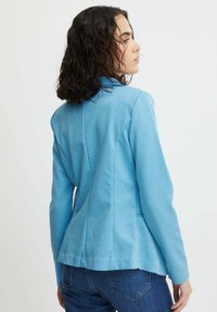 Veste tailleur bleue avec une coupe structurée et un tissu texturé, mettant en avant une taille cintrée et des détails élégants de coutures à l'arrière.