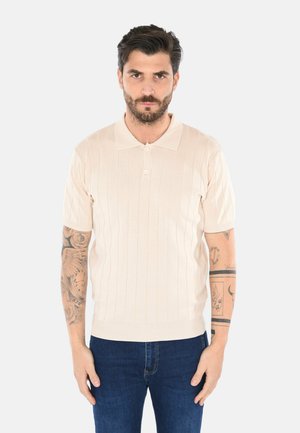 Uomo con capelli scuri e barba, indossa una polo a maniche corte in maglia color crema e jeans blu, in piedi con le braccia rilassate, mostrando i tatuaggi su entrambe le braccia.