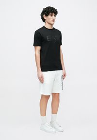 Fiatal férfi göndör hajjal, fekete EA7 pólót, fehér Emporio Armani rövidnadrágot, fehér zoknit és fehér sportcipőt visel, és egyszínű háttér előtt áll.