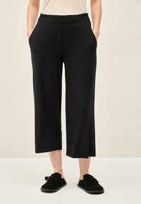 Zwarte, broek tot halverwege de kuit met een elastische tailleband, zijzakken en een gladde textuur. Gecombineerd met zwarte instap schoenen.