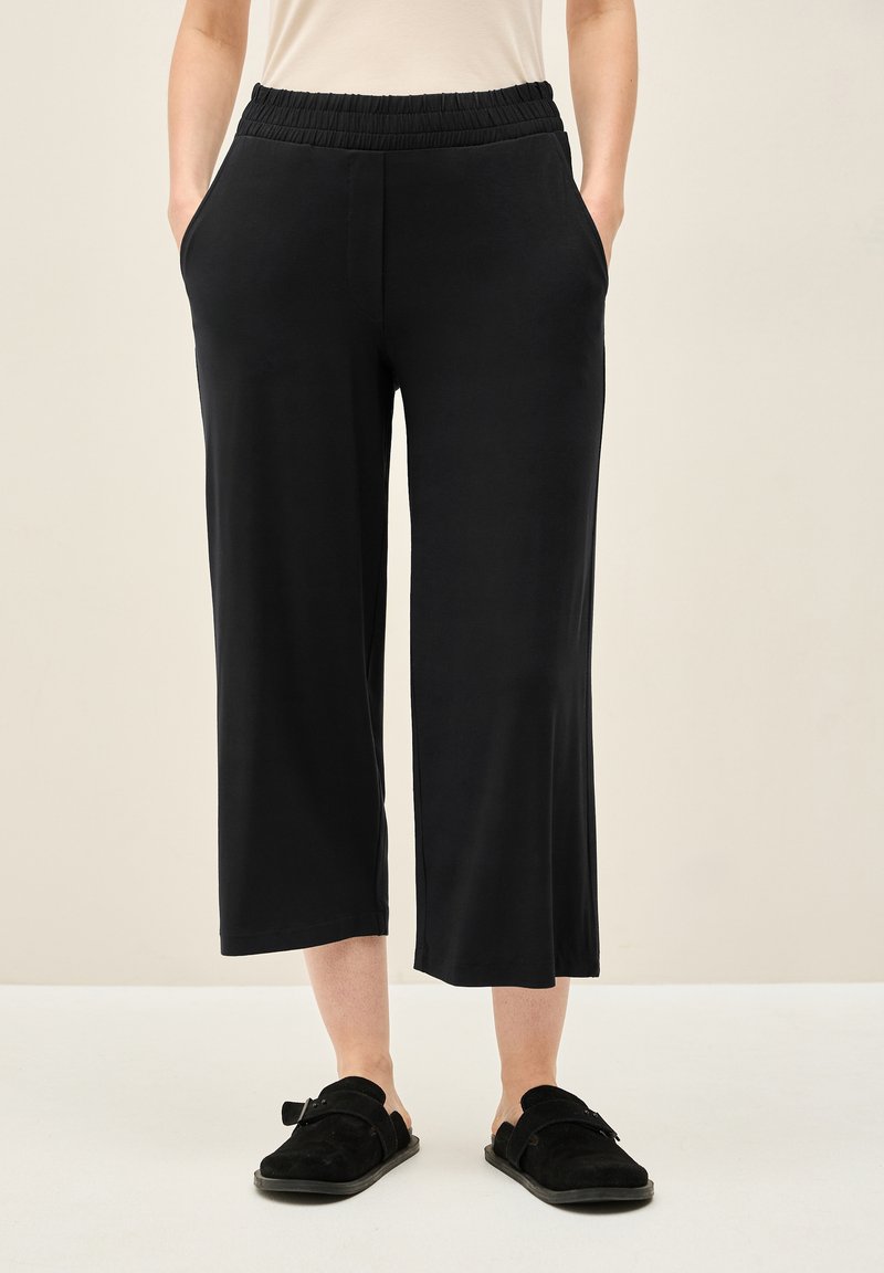 Zwarte, broek tot halverwege de kuit met een elastische tailleband, zijzakken en een gladde textuur. Gecombineerd met zwarte instap schoenen.