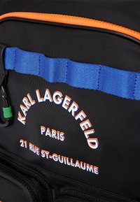 Zaino in nylon nero con cerniera arancione, tracolla blu e dettagli testuali stampati "Karl Lagerfeld", "Parigi" e un indirizzo.