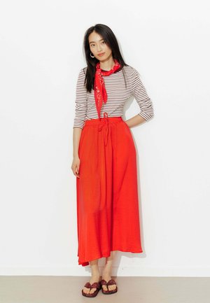 Femme portant un haut rayé blanc et noir, une écharpe rouge, une jupe rouge taille haute et des sandales, debout avec une main sur la hanche.