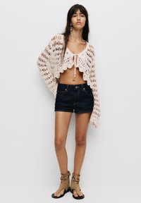 Cardigan din croșetă de culoare bej deschis, cu țesătură deschisă, închidere cu legătură în față, asortat cu șorturi din denim închis și cizme maro deschis cu vârf moale.