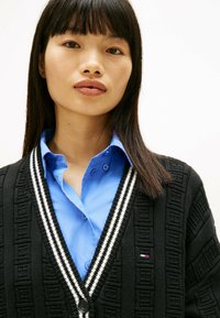 Zwarte cardigan met geometrische textuur, witte gestreepte accenten op de kraag, gedragen over een lichtblauw overhemd met knopen, met een klein logo.