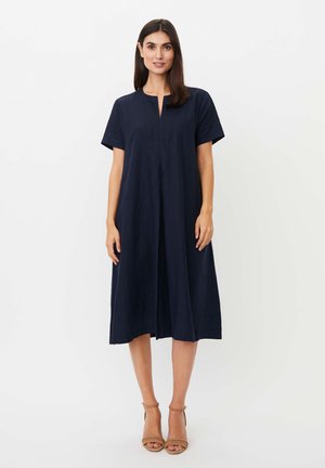 Kobieta stojąca w granatowej sukience midi z krótkim rękawem, z rozcięciem przy dekolcie i beżowych butach na obcasie z paskiem wokół kostki na białym tle.