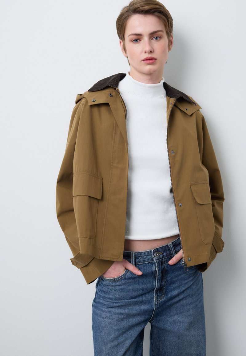 Khaki wasserdichte Jacke mit Reißverschluss, braunem Kragen, zwei aufgesetzten Taschen und leichtem Stoff, kombiniert mit blauen Jeans.