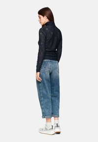 Marinebluse mit langen Ärmeln aus Spitze und geometrischen Mustern, kombiniert mit hellblauen, weiten Jeans. Weiße Sneaker mit schwarzen Akzenten runden das Outfit ab.