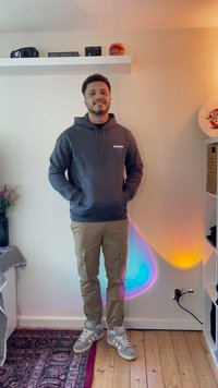 Siva dukserica s logom "dreamer", bež cargo hlače i sivi sneakers. Ima glatku teksturu i ležeran kroj. Šarena ambijentalna rasvjeta u pozadini.