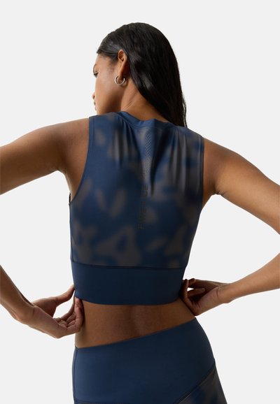 Kvinde justerer navyblå sportstop uden ærmer, synlig ryg og skuldre, med langt mørkt hår og sølvfarvede hoop-øreringe.