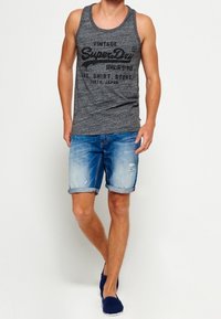 Camiseta sin mangas gris con estampado de "SuperDry", combinada con pantalones cortos de mezclilla azul claro y zapatos náuticos azul marino. Los pantalones cortos tienen puños enrollados y un ligero desgaste.