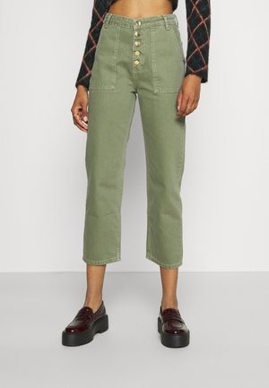 Frau trägt hoch taillierte, kürzere Olivgrüne Hose mit Frontknöpfen, aufgesetzten Taschen, dunkel gemusterten Pullover und schwarze Plateau-Schuhe.