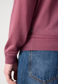 Burgundska dukserica z dolgimi rokavi, izdelana iz mehkega materiala, ima rebrast spodnji rob in manšete; majhna črna oznaka je vidna blizu šiva.