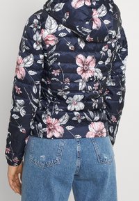 Kukka-aiheinen puffatakki tummansinisessä värissä, jossa on vaaleanpunaisia ja valkoisia kukkia. Suunniteltu hupulla, tikkikuvioinnilla ja kapealla siluetilla.