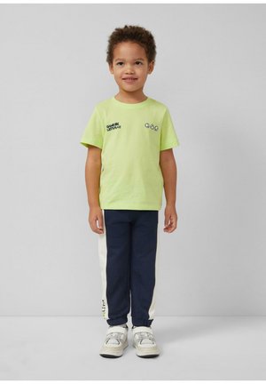 Małe dziecko stojące, ubrane w limonkową koszulkę z napisem "Game on, let's play!", granatowe i białe spodnie oraz białe trampki.