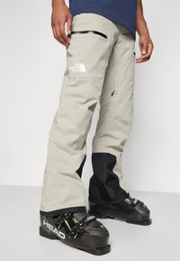 Beige sneeuwbroeken met een zwart onderpaneel, een logo op de dij, ritszakken en aangepast voor compatibiliteit met skischoenen.