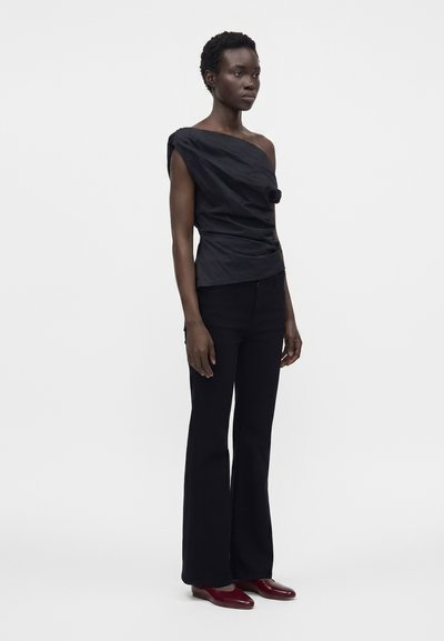 Femme debout portant un top noir à une épaule, un pantalon noir évasé et des chaussures plates bordeaux sur un fond clair uni.