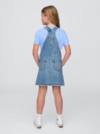 Denim overallkjole med justerbare stropper, lommer og en rå kant. Båret over en lys blå t-skjorte, sammen med hvite høye sneakers.
