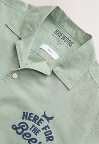 Light green short-sleeved shirt made of textured fabric, featuring a collar, button closure, and navy blue printed text with a bird graphic.