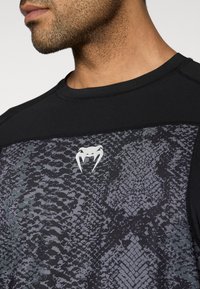 Μαύρο αθλητικό φανελάκι με μοτίβο φολιδωτού δέρματος σε αποχρώσεις του γκρι, με μαύρο λαιμόκοψη και λευκό λογότυπ ο στο κέντρο.