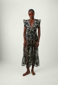 Áttetsző, fekete maxi ruha geometriai mintával, redőzött vállpántos ujjakkal, V-nyakkal és húzózsinóros derékkal, fodros szegéllyel. Narancssárga napszemüveggel viselve.