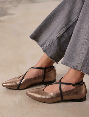 Bailarinas de cobre metálico con puntera afilada, correas negras sobre el pie y un tacón bajo, combinadas con pantalones anchos de mezclilla gris.