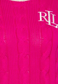 Jasnoróżowy sweter z dzianiny w warkocze, z teksturowanym wzorem i białym haftowanym logo. Projekt zawiera otwarte sekcje dzianiny w całym swetrze.