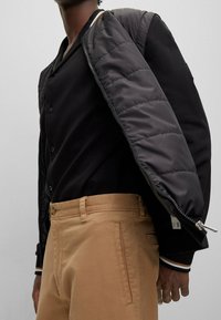 Svart bomberjacka med quiltad svart foder, bärs över en svart knapp-up skjorta, i kombination med khakifärgade byxor med sidfickor.