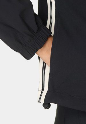 Main partiellement dans la poche d'une veste noire avec bordure à rayures blanches et noires et poignet élastique sur fond blanc.