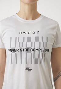 Valge lühikeste varrukatega T-särk, millel on must graafikatekst "HYROX NEVER STOP COMPETING", mustriga aktsentide ja ümarkaeluse disainiga.