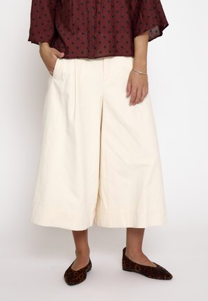 Personne portant un pantalon large crème raccourci, une chemise bordeaux à pois noirs et des chaussures plates à imprimé léopard.