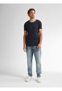 Tricou scurt cu mânecă Army, cu guler rotund, purtat cu blugi de culoare albastru deschis și adidași albi, stând pe un fundal neutru.