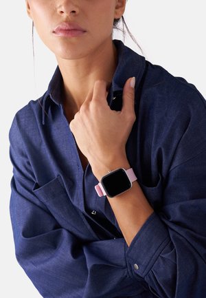 Smartwatch med en firkantet sort urskive, metallhardware og et blødt pink rem. Båret på et håndled, med en blå skjorte i baggrunden.