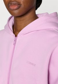 Ροζ φούτερ με φερμουάρ μπροστά, κατασκευασμένο από απαλό ύφασμα, με διακριτικό ανάγλυφο λογότυπο "STRONGER" στο στήθος και κουκούλα με κορδόνι.