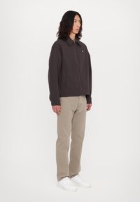 Jachetă zip-up de culoare maro închis cu guler, buzunare laterale, purtată peste pantaloni bej deschis și adidași albi. Textură netedă în general.