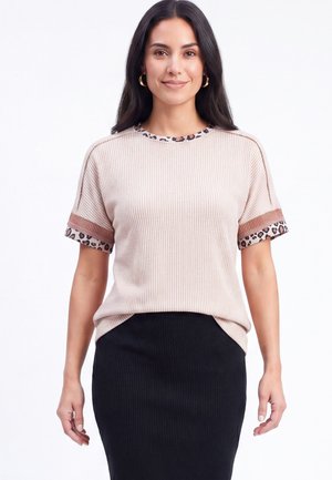 Kvinde med langt mørkt hår iført beige kortærmet ribbet top med leopardprintkant og sort ribbet nederdel, stående mod hvid baggrund.