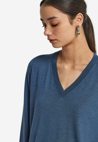 Niebieski, dziany sweter w serek z długimi rękawami. Gładka tekstura i minimalne detale. Modelka nosi asymetryczny srebrny kolczyk.