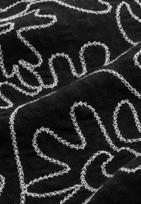 Gros plan sur un tissu noir avec une broderie zigzag blanche formant des motifs et formes abstraites.