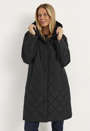 Woman wearing a long black quilted coat with a hood, holding the hood edges while looking slightly to the side and smiling.