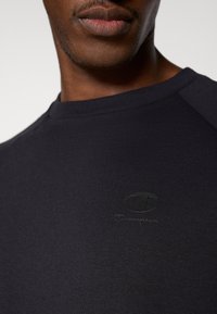 Σκούρο μπλε μπλουζάκι από απαλό, λείο ύφασμα με στρογγυλή λαιμόκοψη και μανίκια ράγκλαν. Διαθέτει διακριτικό μαύ ρο λογότυπο Champion στο στήθος.