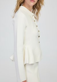 Chaqueta peplum blanca con un cuello estructurado, mangas largas y detalles de botones dorados. Tejido texturizado con una silueta ajustada.