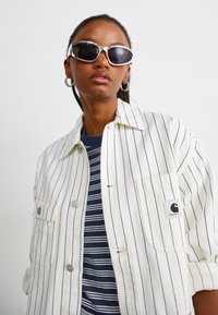 Veste à rayures blanches et marines avec deux poches avant, des fermetures à bouton et un patch logo, superposée sur une chemise à rayures marines et blanches. Lunettes de soleil noires.