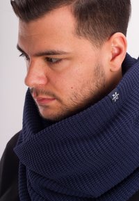 Navy knitted scarf with a thick, textured pattern. Features a small logo tag in the corner, wrapping closely around the neck.