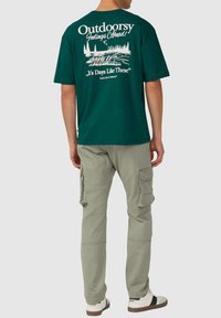 Groene katoenen T-shirt met een print van bomen en water op de achterkant. Gecombineerd met lichtgrijze cargobroeken en casual witte sneakers.