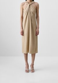 Robe midi beige sans manches avec un décolleté licou torsadé portée avec des sandales blanches à talons hauts et brides sur un fond gris.