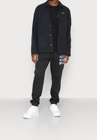 Svart jacka med två framfickor, svart t-shirt, svarta byxor med vit "NIKE"-text och vita träningsskor.