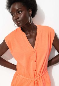 Robе en orange sans manches avec un décolleté en V, des boutons sur le devant et une taille froncée. Le tissu semble doux avec une texture légère.