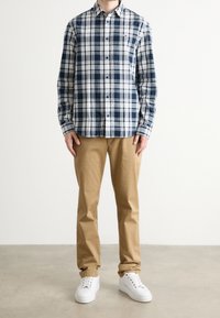 Osoba ubrana w granatowo-białą koszulę w kratę zapinaną na guziki, beżowe spodnie i białe trampki, stojąca na szarej podłodze na tle jednolitej białej ściany.