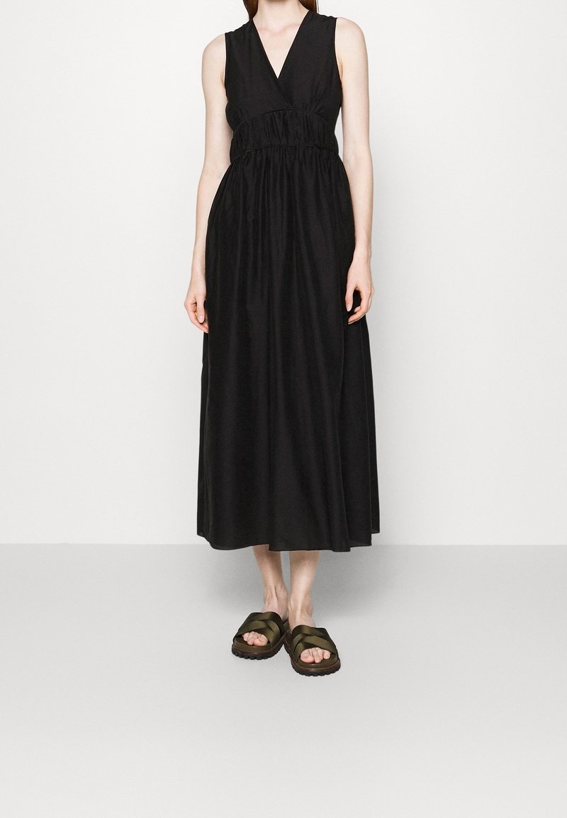 Femme en robe midi noire sans manches avec un décolleté en V profond et une taille froncée, portant des sandales à brides croisées vert olive.